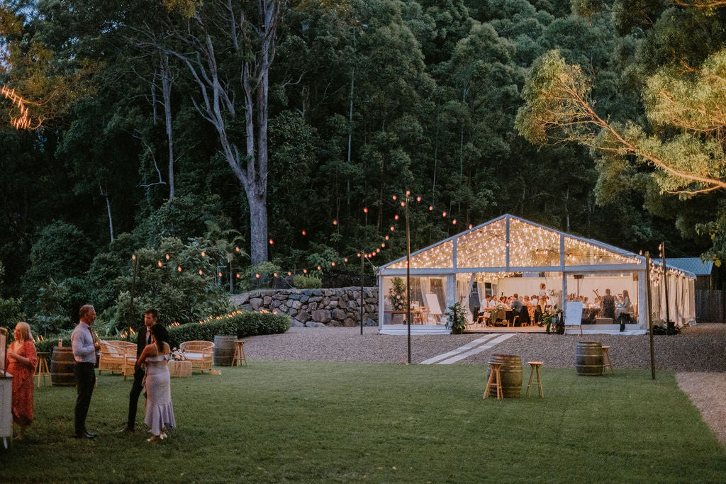 Afternoon view of a wedding venue with a clear marquee, surrounded by green trees, with people enjoying the place.